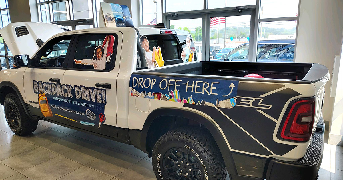 A white pickup truck in a showroom with "Backpack Drive" graphics, featuring smiling kids and school supplies. Sign reads "Drop Off Here," conveying a charitable tone.